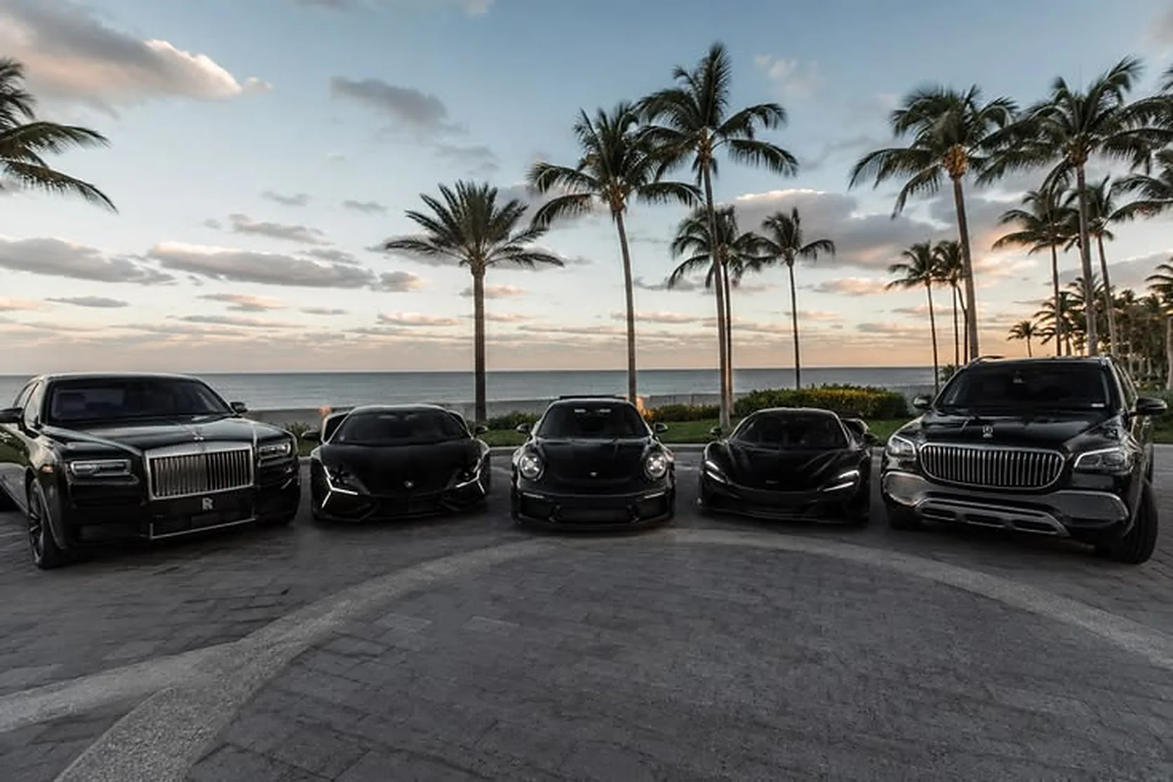 Rolls-Royce, Lamborghini, Porsche, McLaren y Mercedes-Maybach negros alineados junto al mar al atardecer en Dubái